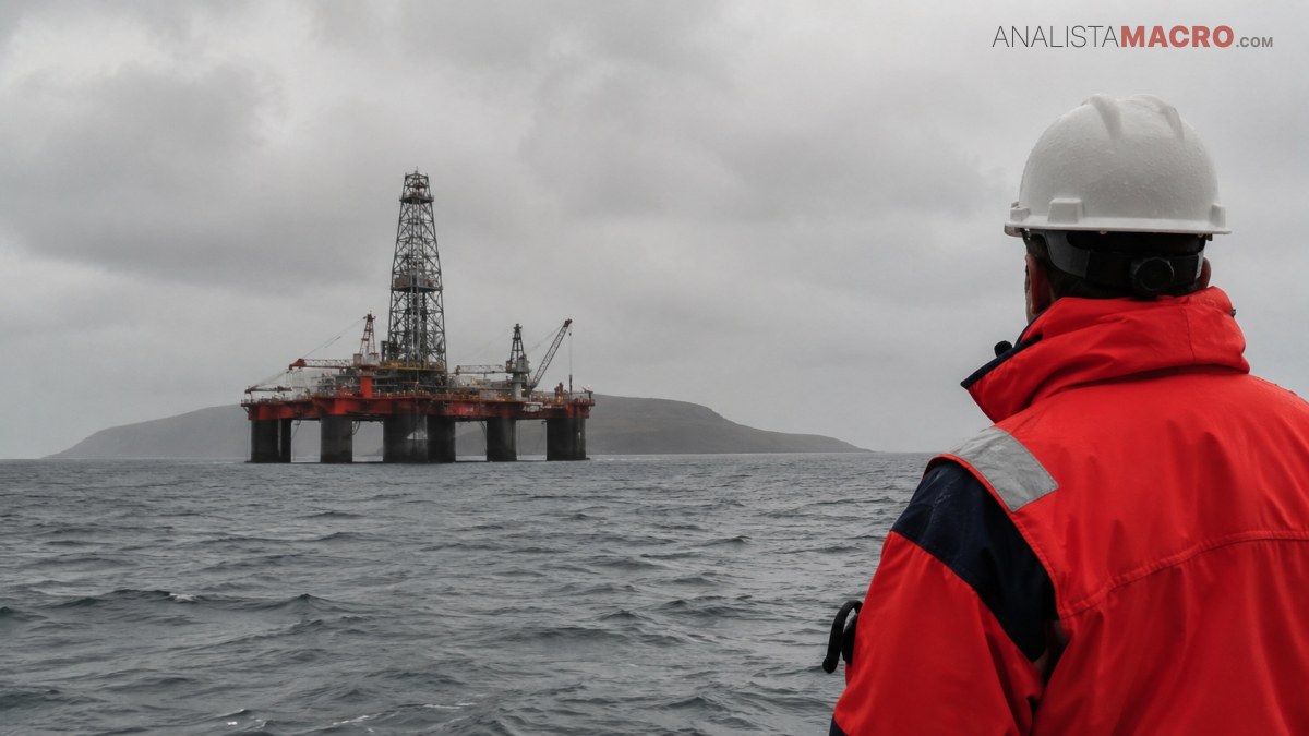 Persona con casco blanco y chaleco rojo observa una plataforma petrolera sobre el mar agitado, bajo un cielo nublado.