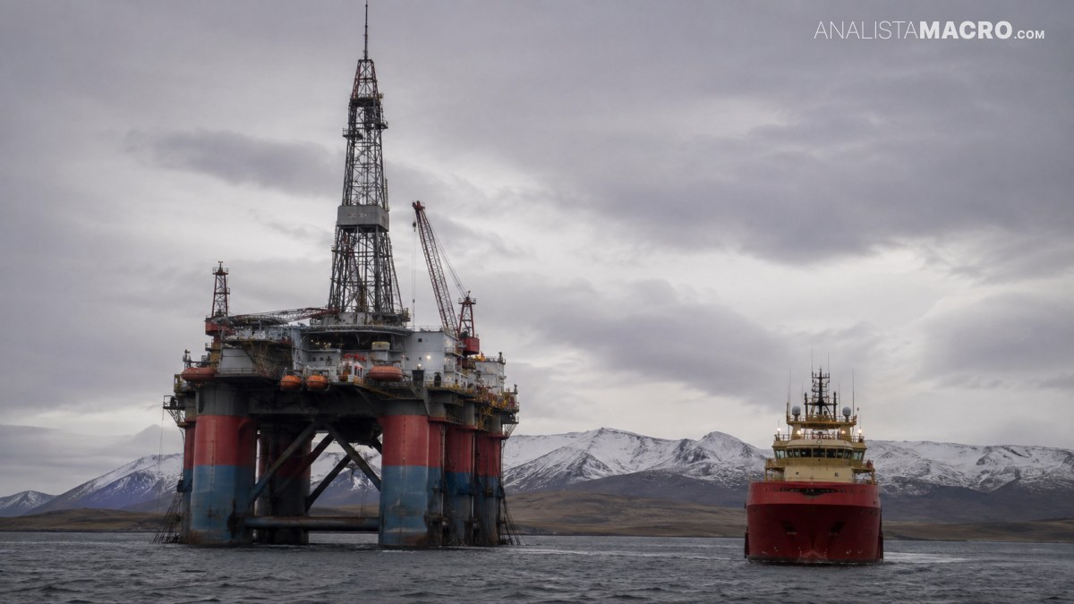 Plataforma petrolera marina junto a un barco rojo, con montañas nevadas y cielo nublado al fondo.