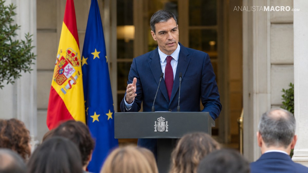 Hombre con traje habla ante un atril, frente a banderas de España y la Unión Europea, mientras varias personas lo observan.