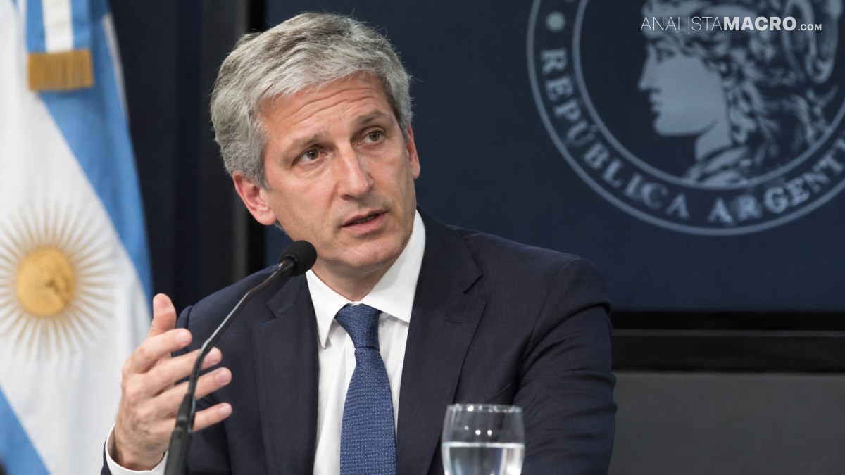 Hombre de cabello canoso, traje oscuro y corbata, gesticulando frente a un micrófono con una bandera al fondo.