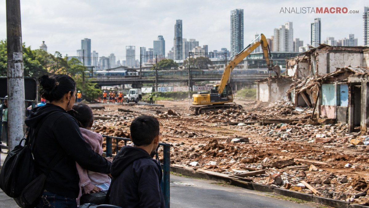 Una mujer con dos niños observa una zona de demolición con una excavadora, escombros y edificios altos al fondo.