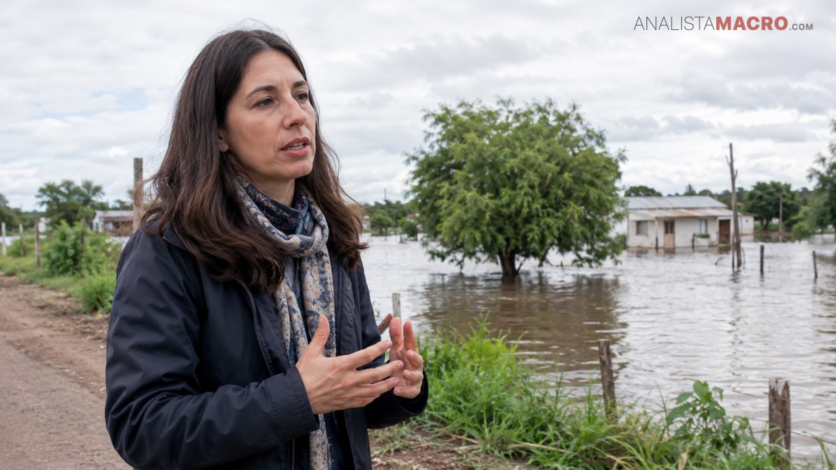 Mujer de cabello oscuro habla junto a una zona inundada, con árboles, viviendas y agua acumulada al fondo.