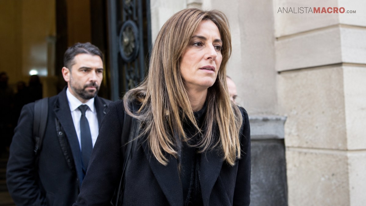 Mujer de cabello largo castaño y abrigo negro camina frente a un edificio, seguida por un hombre vestido formalmente.