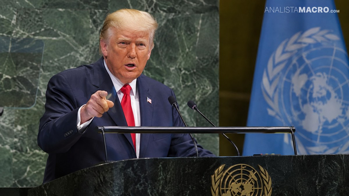Donald Trump habla ante un podio, señalando hacia el frente, con una bandera de las Naciones Unidas al fondo.