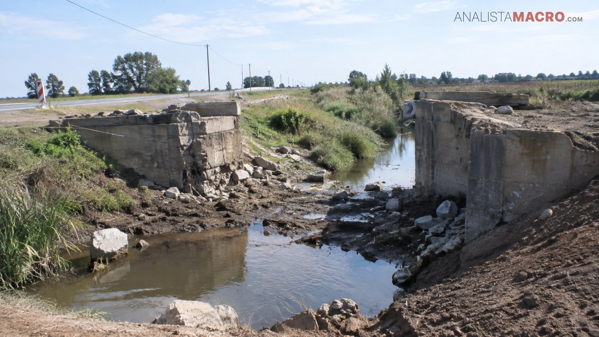 Canal de agua atraviesa un puente de hormigón deteriorado, rodeado de vegetación, campos y árboles bajo un cielo despejado.