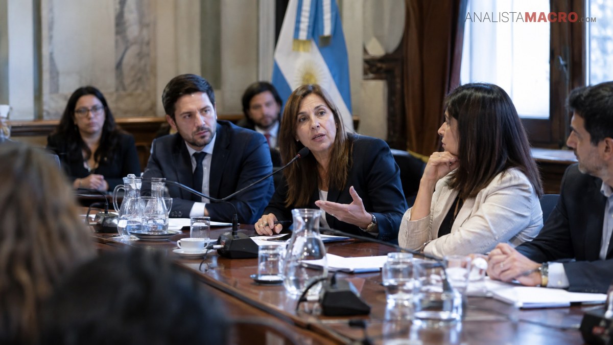 Personas participan en una reunión alrededor de una mesa, con documentos, vasos y micrófonos, mientras una mujer habla.